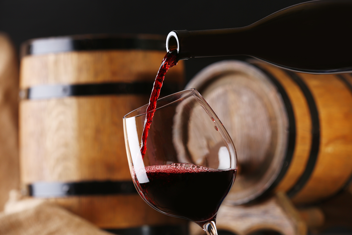 Pouring red wine from bottle into glass with wooden wine casks on background